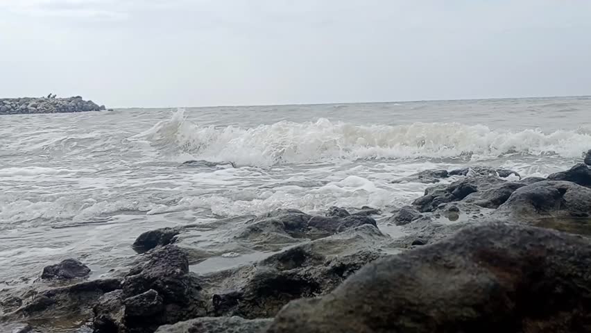 Footage of a natural rocky beach scene with white waves crashing against black coral. A cloudy sky creates a calm, cool, and peaceful seascape