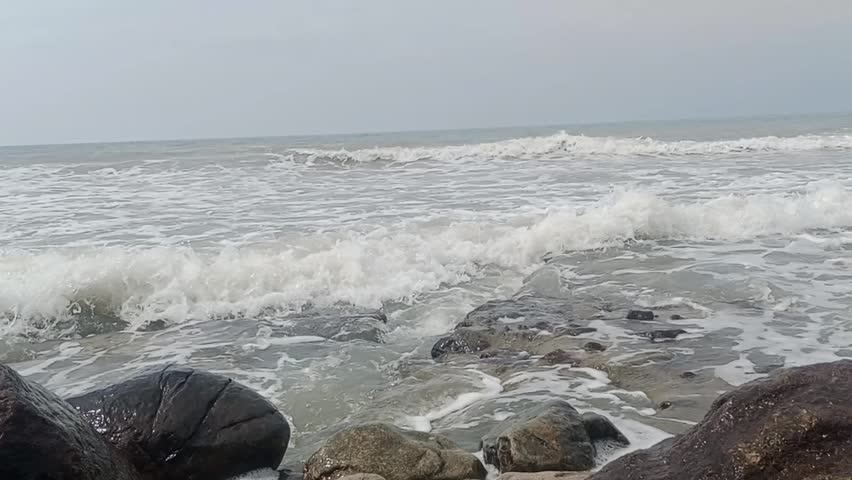 Footage of a natural rocky beach scene with white waves crashing against black coral. A cloudy sky creates a calm, cool, and peaceful seascape