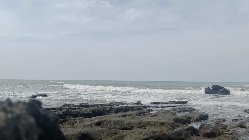 Footage of a natural rocky beach scene with white waves crashing against black coral. A cloudy sky creates a calm, cool, and peaceful seascape