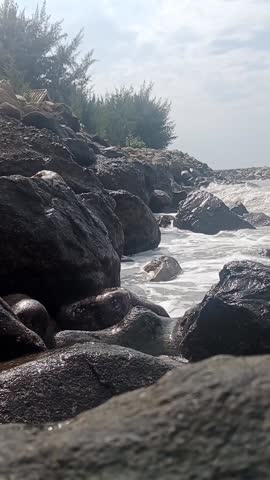 Footage of a natural rocky beach scene with white waves crashing against black coral. A cloudy sky creates a calm, cool, and peaceful seascape