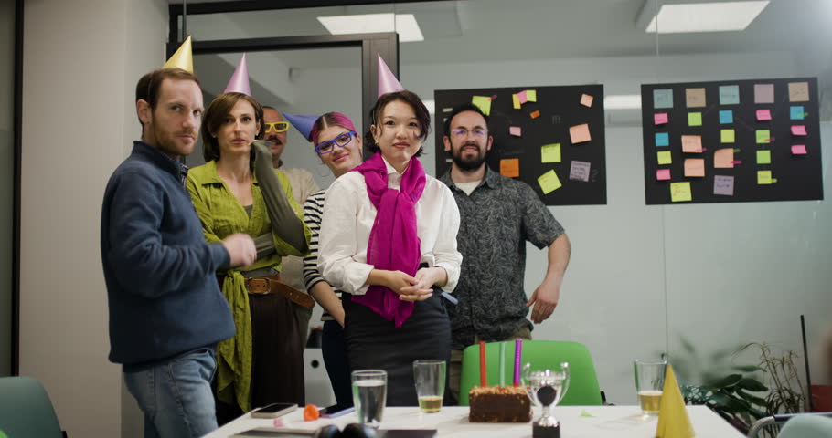 A group of five people gathers in an office. They cheerfully raise their hands and enjoy cake at a table. The mood is lively as they celebrate a work achievement in the afternoon.