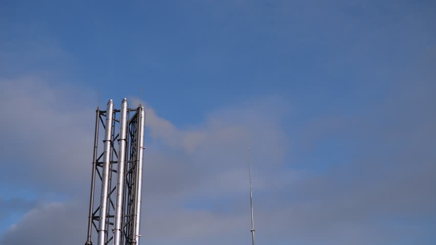 industrial steel tower against blue sky, tall metal lattice mast with antenna, calm daylight and scattered clouds, urban infrastructure silhouette, maintenance ladder and platform visible,