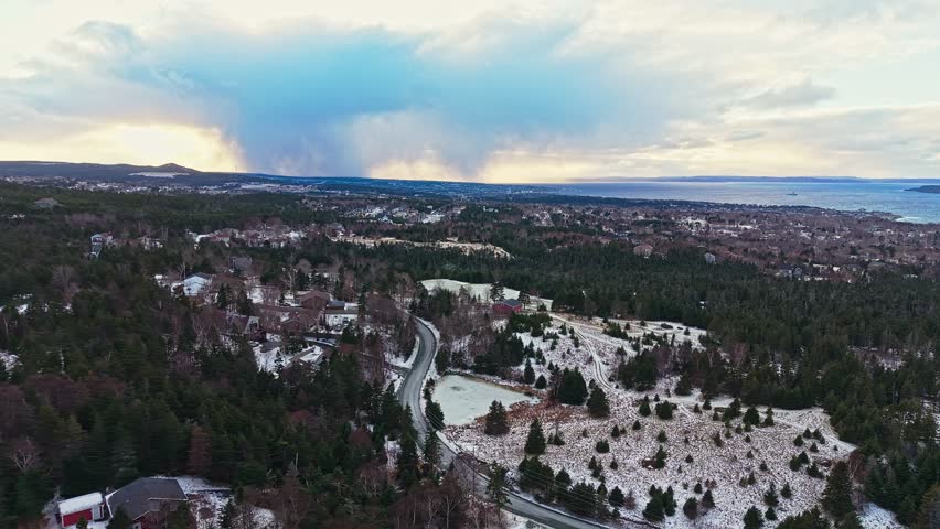 Aerial drone shot sweeps over a snow-speckled forest and winding road through a rural landscape with dramatic clouds looming on the horizon.