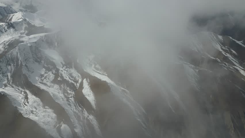 Aerial view captures the stunning snow-capped mountains partially obscured by clouds, showcasing the rugged terrain and dramatic textures, as the camera pans slowly across the landscape
