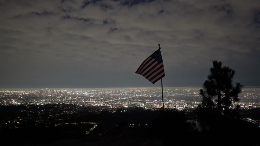 Camera shot of a United States flag waving at night with Los Angeles cityview in the background