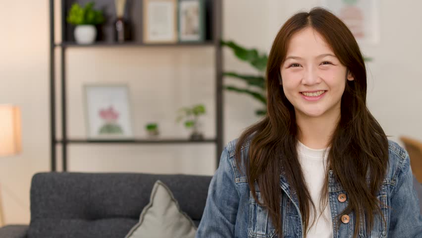 Cheerful young woman in denim jacket smiles confidently on sofa in bright indoor living room