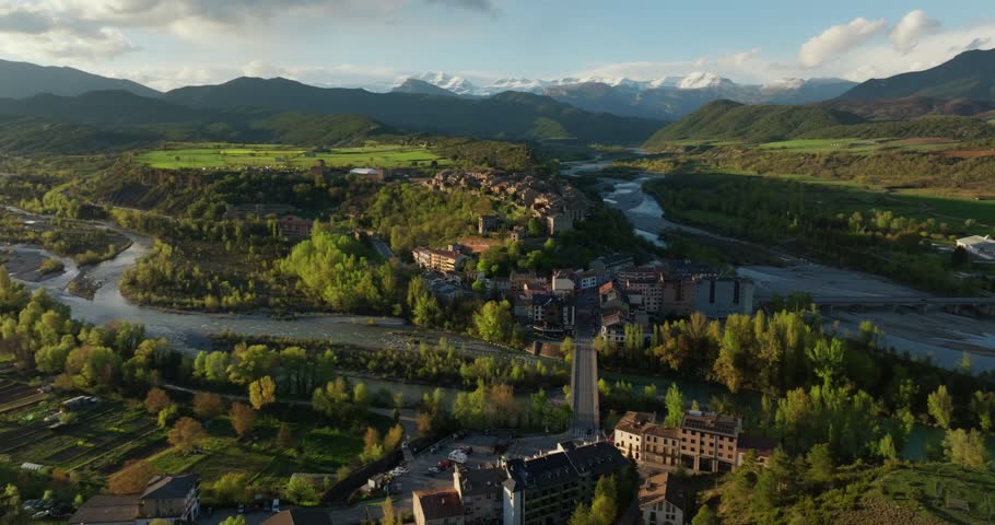 Drone glide approaches Aínsa from the Mirador de Fuebla side, flying above the Río Cinca channels, riverside woodland toward fortified medieval village and castle on hill with snowy Pyrenees peaks.
