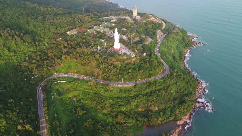 Top-down aerial view of the Lady Buddha statue and Linh Ung Pagoda complex on Son Tra Peninsula during golden hour. Shows the winding coastal road and ocean in Da Nang, Vietnam