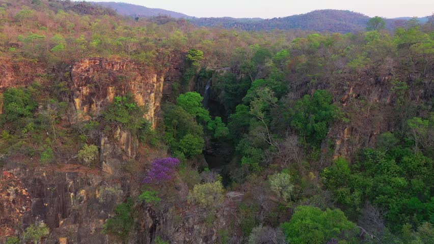 Drone advances forward into Cerrado canyon approaching waterfall surrounded by rocky cliffs and lush riparian vegetation corridor in protected reserve Goiás state Brazil.