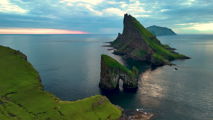 Witness the stunning Drangarnir cliffs in the Faroe Islands as they rise from the Atlantic Ocean. The serene landscape is illuminated by the fading sunlight, creating a magical atmosphere.