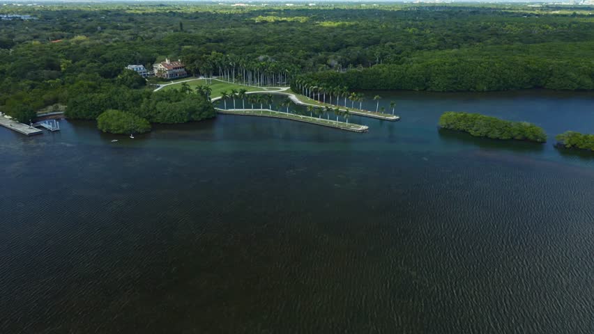 Calm water body bordered by dense green forest and a waterfront estate with palm trees. The tranquil scene captures natural beauty with a blend of lush vegetation and clear water.
