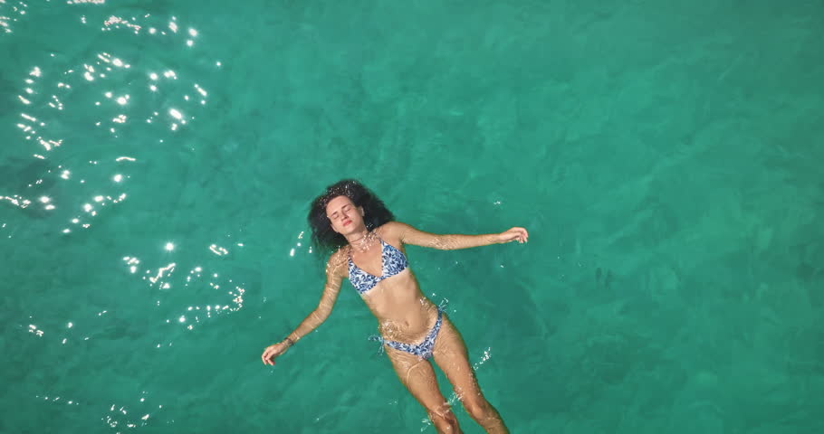 Young woman in bikini relaxing floating on back in crystal clear turquoise ocean water, enjoy serene summer holidays and vacation in tropical paradise. Aerial top down zoom out panorama drone flight