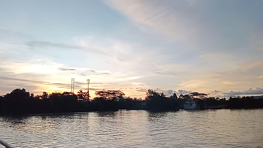 Wide shot of a tropical river at sunset with silhouettes of trees and distant mountains under a dramatic cloudy sky. Calm water with soft ripples reflecting the fading evening light.