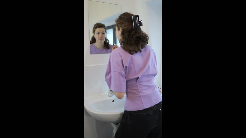 Woman in bathroom applies makeup looks in mirror and washes her face over white sink during the morning