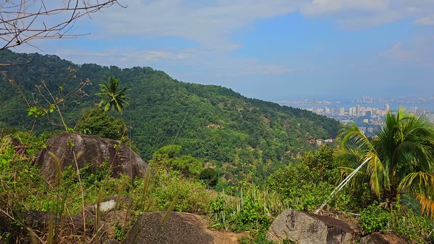 Panoramic view of Penang city and George Town from Penang Hill viewpoint. Urban landscape with high-rise buildings, mountains and tropical greenery under blue sky with clouds. Scenic overlook of island cityscape.