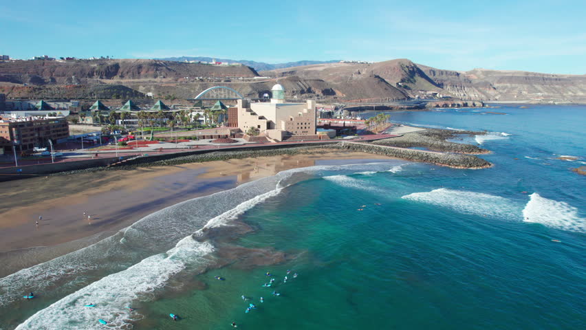 Scenic aerial footage of Las Palmas coastline with surfers riding waves on a bright sunny day. Gran Canaria
