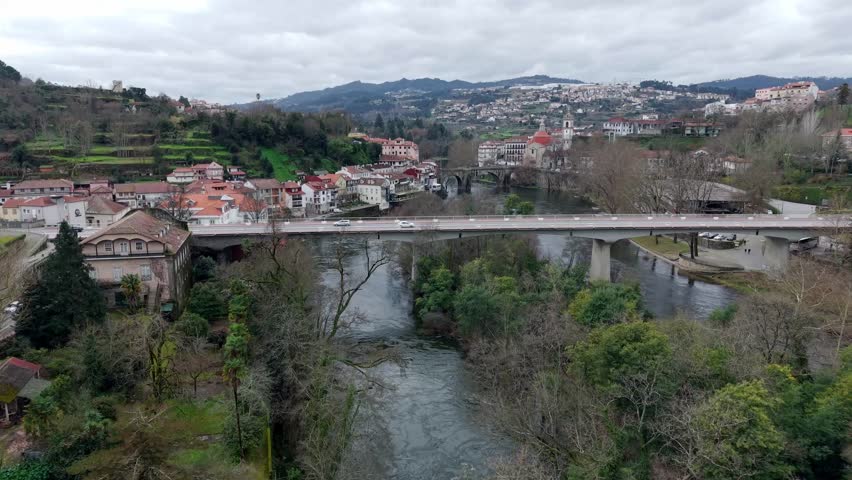New bridge in Amarante city on Tamega riverbank featuring dense mixed autumn forest, Igreja de São Gonçalo catholic church and historic stone bridge, Tracking drone shot