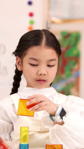 Focused schoolgirl carefully stacking translucent geometry blocks in bright classroom, static medium shot, soft lighting