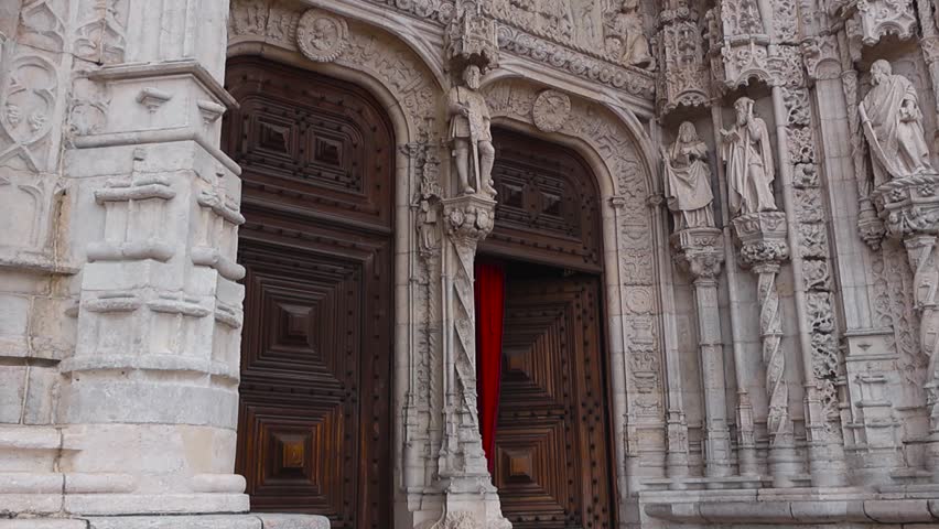 Cinematic upward reveal of the Jerónimos Monastery entrance showing carved stone sculptures and open wooden doors with a red curtain inside at a famous Lisbon landmark.