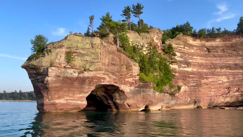 A boat idles on calm water near a unique, rugged rock structure that protrudes from an island. The sand stone features stripes of red and yellow stone, with sporadic pine trees on the rock face. There is a small cave formed by the rock structure. The vibrant colors of the stone stand in stark contrast to the blue hues of the still water and clear, sunny sky. 