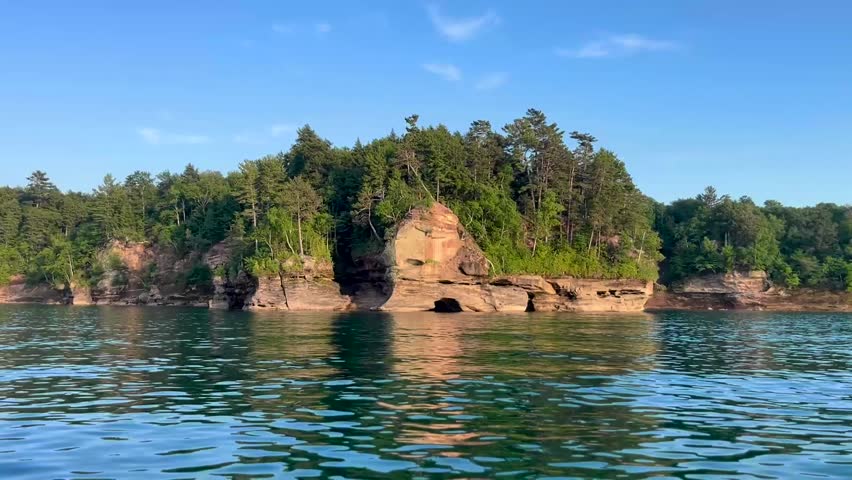 A boat glides across the clear, blue waters of a lake beneath a sunny, blue sky. The boat passes by a rugged rock face shoreline topped with a lush evergreen forest. This scenic landscape view captures a relaxing boat view of a unique geologic structure in the Upper Peninsula of Michigan. 