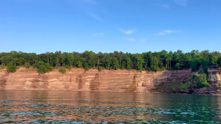 A boat glides across the clear, blue waters of a lake beneath a sunny, blue sky. The boat passes by a rugged rock face shoreline topped with a lush evergreen forest. This scenic landscape view captures a relaxing boat view of a unique geologic structure in the Upper Peninsula of Michigan. 