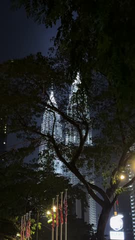 Artistic vertical night shot for social media content. This handheld video captures a unique, cinematic approach to the iconic Petronas Twin Towers in Kuala Lumpur. Filmed through the dense green foliage of trees, the illuminated skyscrapers shine with a bright white glow against the deep black night sky, creating a mysterious and atmospheric frame.
The camera moves steadily forward, simulating a person's walk towards the towers. This POV shot adds dynamism and a sense of discovery, perfect for travel vlogs, intro sequences, or transitions about adventure, city life, or hidden perspectives. In the foreground, motionless flagpoles add geometric lines to the organic shapes of the leaves, emphasizing the windless, still night.
This clip is ideal for projects requiring authentic, artistic, and visually striking footage of famous landmarks from an unusual angle. Perfect for social media posts - Reels, Shorts, Stories, - travel advertising, website backgrounds, or any project needing a touch of cinematic mystery and urban exploration.