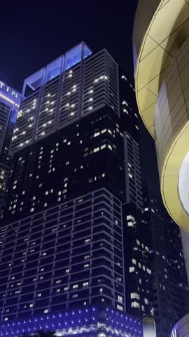 A seamless, loopable vertical cinematic video perfect for social media and advertising. This shot captures the essence of a modern, luxurious metropolis at night. It begins with a meditative focus on the intricate details and flowing water of an architectural fountain, the primary soothing sound in the scene.

The camera then executes a smooth, continuous movement, tilting up along the majestic, illuminated glass facade of a skyscraper. In its dark windows, a reflection of the vividly lit KL Tower is visible, adding a layer of iconic depth. The motion continues upward into the starless night sky, pans across the tops of neighboring high-rises, and gracefully descends back to earth, revealing the lush greenery of palm trees and the sophisticated ground-level lighting of the urban landscape.

This clip is ideal for promoting high-end real estate developments, luxury business districts, urban design projects, and city tourism. Its serene, people-free composition and elegant pacing make it suitable for corporate presentations, website backgrounds, and social media content - Reels, Shorts, Stories - focusing on architecture, economic growth, and sophisticated urban living.