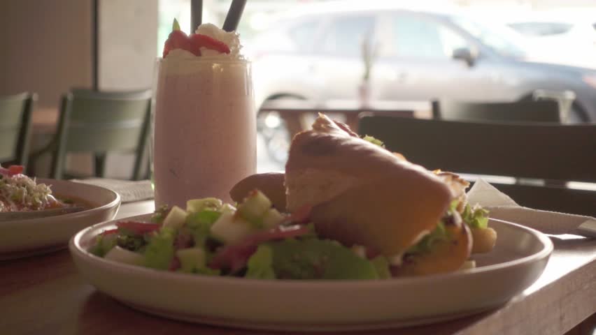 A complete meal on a restaurant table. Features a fresh baguette sandwich, a green side salad, and a pink milkshake in the background near a window.
