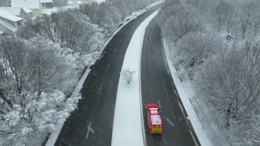Aerial tracking shot of winter road in United States, with snow-covered trees and roadside while a emergency firetruck vehicle drives along cleared highway. Calm and cold American winter scene.