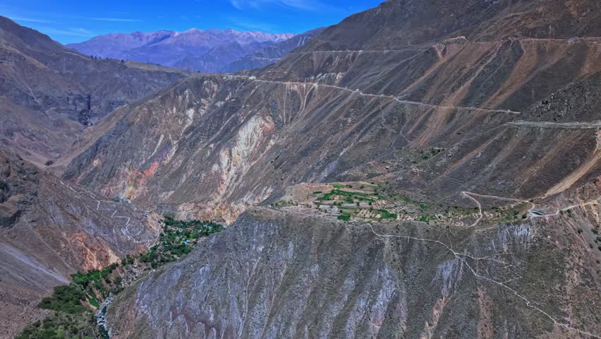 Drone footage looking straight down center of Colca Canyon in Peru. The lush oasis and farmland sit below while dramatic canyon walls, distant mountains, and patches of blue sky create depth and scale