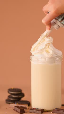 Close-up of hand swirling whipped cream onto vanilla milkshake, warm studio lighting, static camera
