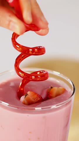 Hand inserts and twirls red spiral straw in creamy strawberry smoothie with fresh fruit garnish