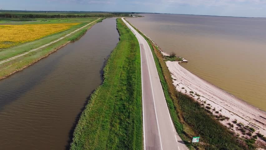 Drone view of a winding road curving along a lake and canal, surrounded by green farmland and distant horizon in daylight.