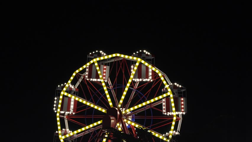 A brightly lit Ferris wheel with yellow and white bulbs spinning against a pitch-black sky at an amusement park or carnival fairground.