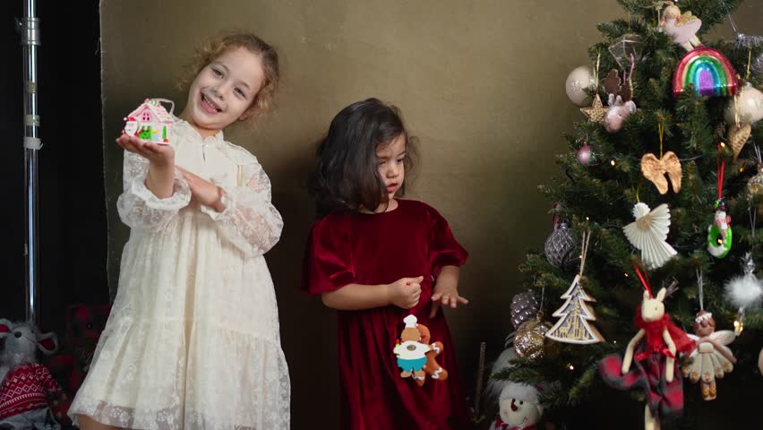 A little girl in a white dress smiles while holding up a pink house decoration next to a festive Christmas tree and her sister