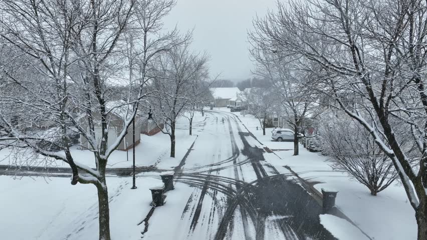 Aerial Forward flight through quiet American suburban neighborhood after snowfall, with tree branches framing snow-covered houses and tire tracks leading along calm residential street. Wide shot.