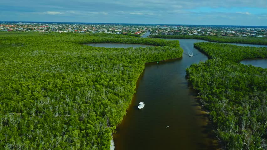 A white boat travels on a narrow river surrounded by lush green mangroves, under a cloudy sky with a housing development visible far in the background.
