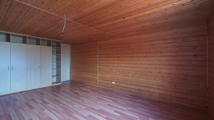 Spacious room with wooden paneling on walls and ceiling, wooden flooring, large white wardrobe with glass inserts, white radiator, minimal furnishing, warm natural tones.