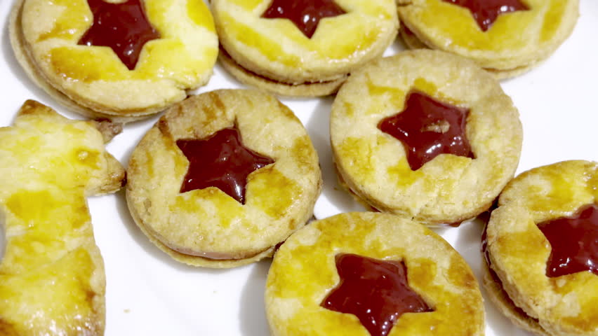 Close-up of homemade Alsatian bredele sandwich cookies with star-shaped red jam filling, golden egg-wash glaze on white plate, festive Christmas holiday dessert