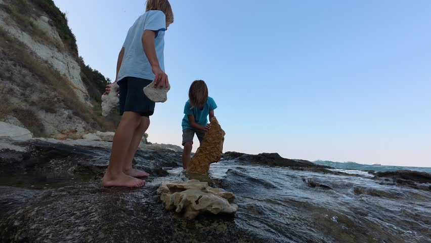 Two children playing with stones on rocky seashore as waves splash around. Summer coastal adventure with exploration, nature play and joyful childhood by the sea