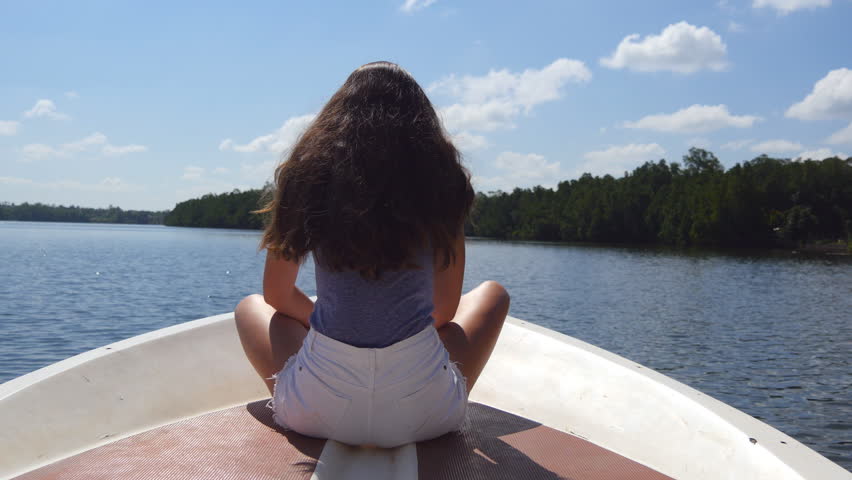 Unrecognizable girl with blowing long hair sits on bow of boat and admires a beautiful nature landscape. Young woman rests on deck of yacht and enjoys summer travel. Concept of vacation or holiday