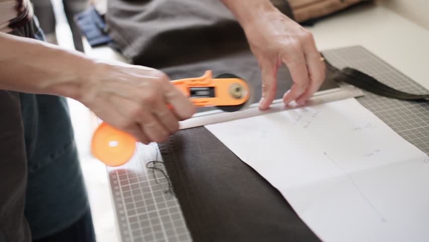 Tailor measuring and cutting black fabric with rotary cutter and ruler while working on garment design pattern in workshop