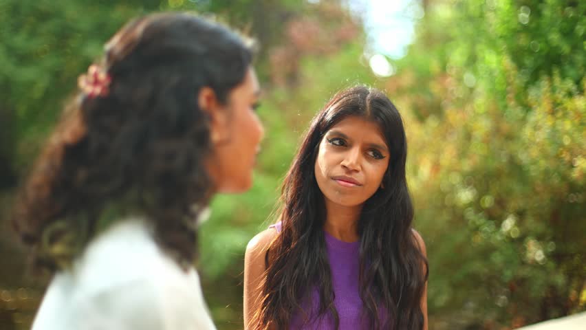 Lovely young indian woman with long hair listening to her friend while talking outdoors in a park, her expression changing as she listens