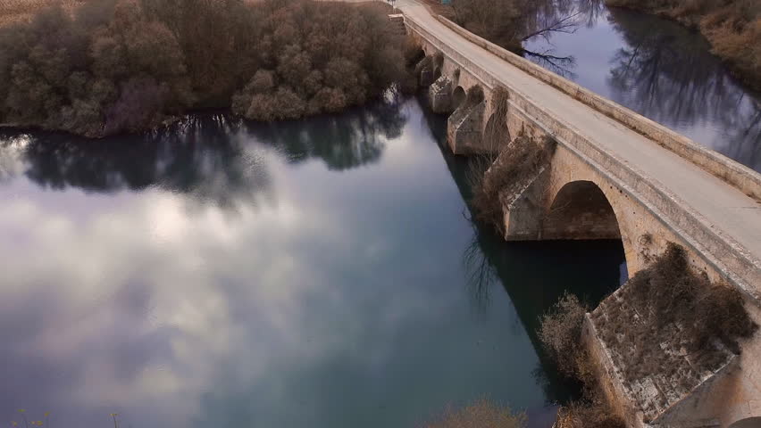 Aerial drone video flying backward from Fitero bridge in Palencia Spain, crossing the Pisuerga River. Smooth pull back shot revealing stone arches, reflections, calm water, and autumn landscape. 