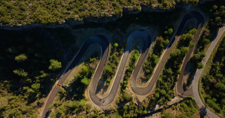 Wide overhead drone static shot reveals cyclists threading steep hairpin switchbacks across a forested mountain overlooking Cinca River gorge in Sobrarbe, with multiple curves visible