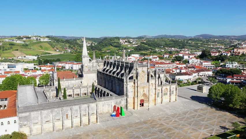 Drone shot of the historic Batalha Monastery surrounded by the town and green hills under a clear blue sky in Batalha, Portugal.