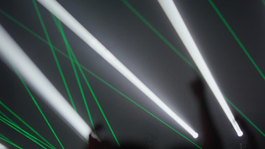 Green and purple laser beams cross above cheering crowd during live music show with hands raised in excitement