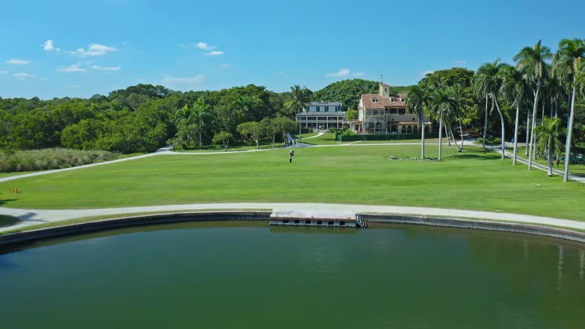 Historic estate with expansive green lawns, tropical palm trees, and a tranquil pond featuring a dock. The clear sky and surrounding forest add to the peaceful atmosphere of the location.