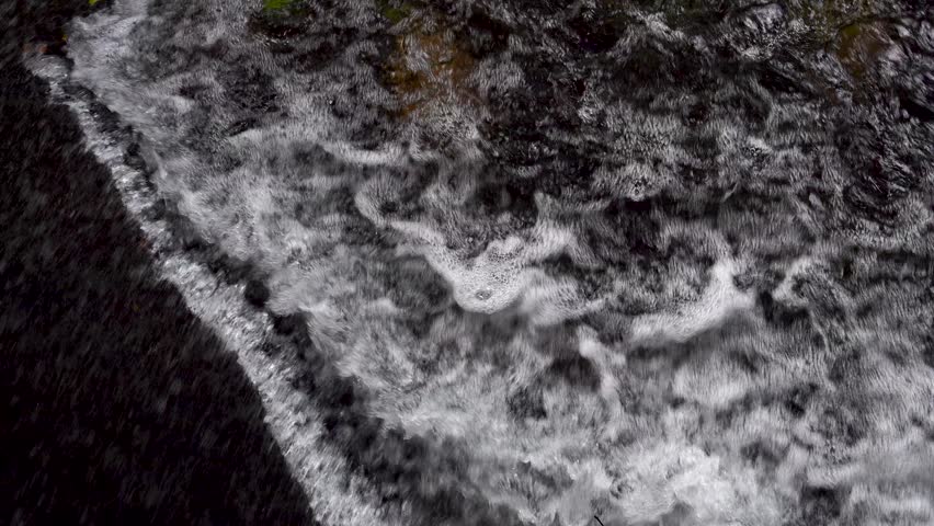 Panning overhead view of turbulent river water surface flowing rapidly over a dam. Rushing stream creating white foam and bubbles. Concept of hydro energy, power, and water management.